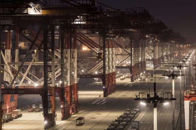 © Bloomberg. Trucks drive past gantry cranes at the Yangshan Deepwater Port, operated by Shanghai International Port Group Co. (SIPG), in the early morning in Shanghai, China, on Wednesday, Aug. 7, 2019. Trump's threat to raise tariffs on all Chinese goods last week shattered a truce reached with Xi just weeks earlier, unleashing tit-for-tat actions on trade and currency policy that risk accelerating a wider geopolitical fight between the world's biggest economies. Photographer: Qilai Shen/Bloomberg