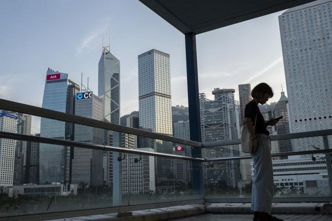 © Bloomberg. A pedestrian using a smartphone is silhouetted as the Bank of America Tower, from left, AIA Central building, CCB Tower, Cheung Kong Center building, HSBC Holdings Plc headquarters building, Standard Chartered Bank building, Jardines House building and other buildings stand in the background in the Central district of Hong Kong, China, on Sunday, Nov. 3, 2019. The record weakness facing Hong Kong's hard-hit retailers continued, albeit at a slower pace, in September as violent protests helped push the city into a recession, data released on Nov. 1 showed. Photographer: Chan Long Hei/Bloomberg