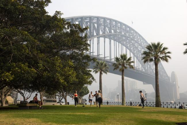 © Bloomberg. People stretch at a park as the Sydney Harbour Bridge is shrouded in haze in Sydney, New South Wales, Australia, on Wednesday, Jan. 8, 2020. As Sydney faced another day of toxic haze shrouding the skyline, U.S. weather satellite captured the smoke crossing South America and spreading out over Buenos Aires before it drifted into the Atlantic Ocean -- some 7,328 miles (11,793 kilometers) east of Sydney -- according to the National Oceanic and Atmospheric Administration. Photographer: Brent Lewin/Bloomberg