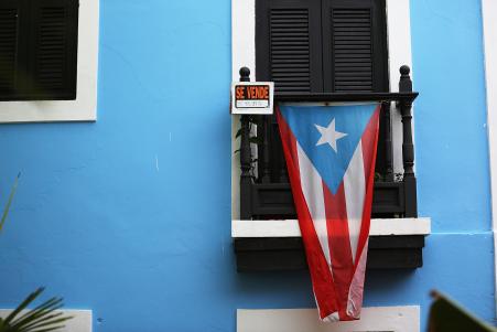 © Joe Raedle/Getty Images. A for sale sign is seen hanging from a balcony next to a Puerto Rican flag in Old San Juan as the island's residents deal with the government's  billion debt on July 1, 2015 in San Juan, Puerto Rico. Banks enabled the U.S. commonwealth's political leaders to delay addressing needed reforms for years, earning fees along the way and passing the risk to bond buyers.