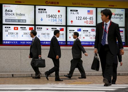 © Reuters. Businessmen walk past screens displaying market indices outside a brokerage in Tokyo, Nov. 16, 2015.