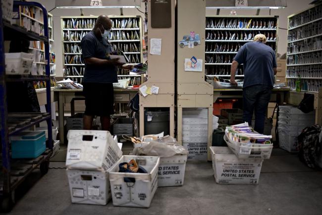 © Bloomberg. A letter carrier wears a protective mask while preparing mail for delivery at a United States Postal Service (USPS) facility in Fairfax, Virginia, U.S., on Tuesday, May 19, 2020. The Postal Service in recent weeks has sought bids from consulting firms to reassess what the agency charges companies such as Amazon, UPS and FedEx to deliver products on their behalf between a post office and a customer's home, the Washington Post reported last week. Photographer: Andrew Harrer/Bloomberg