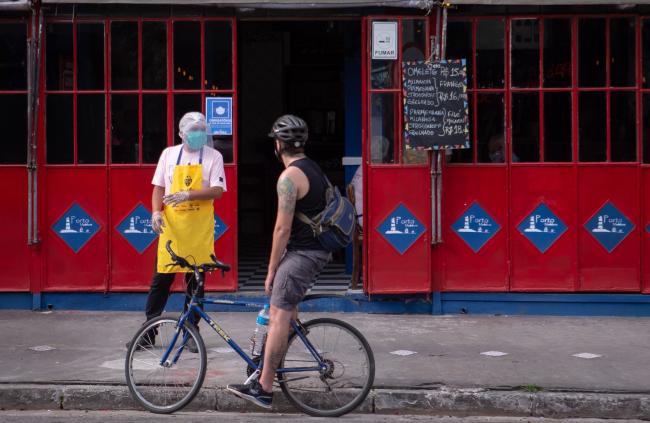 © Bloomberg. A server wearing a protective mask and face shield talks to a cyclist outside a restaurant in the Vila Madalena neighborhood of Sao Paulo, Brazil, on Monday, July 6, 2020. The mega-city, with some 20 million people in the greater metropolitan area, started reopening restaurants and bars Monday even as the virus explodes across much of the country.
