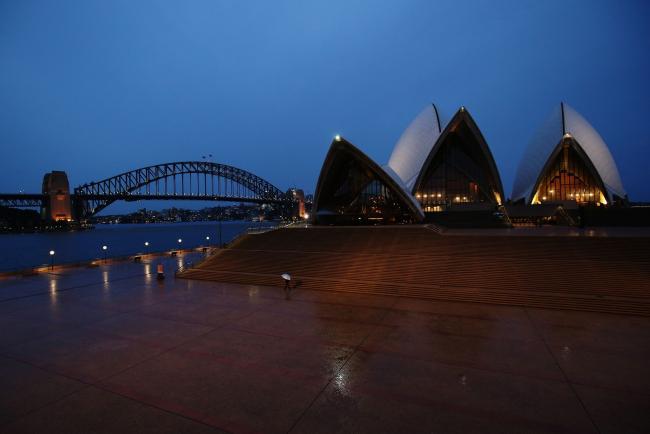 © Bloomberg. A pedestrian holding an umbrella walks along the Sydney Opera House forecourt during a partial lockdown imposed due to the coronavirus, at night in Sydney, Australia, on Friday, April 3, 2020. Australia's Prime Minister Scott Morrison said the government is close to announcing an agreement that will see rent relief for businesses hit by the coronavirus outbreak. The government wants a new industry code of practice for commercial tenancies, so any decline in revenue for a small- or medium-sized enterprise is reflected in a proportionate drop in rent they have to pay. Photographer: Brendon Thorne/Bloomberg