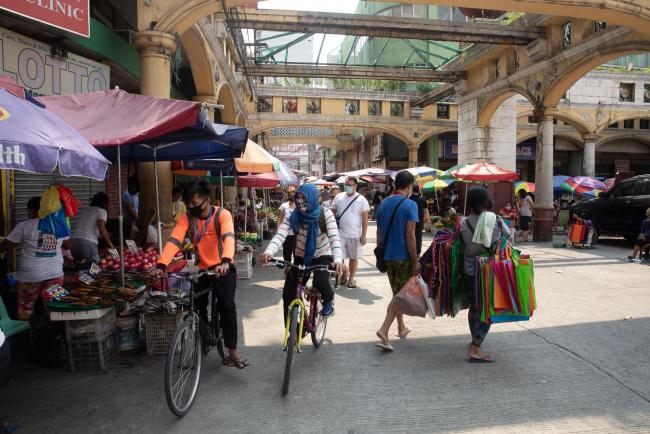 © Bloomberg. Cyclists wearing protective masks ride past stores and shoppers at Quiapo Market in Manila, the Philippines, on Monday, July 27, 2020. President Rodrigo Duterte pushed for corporate tax cuts and targeted support for pandemic-hit industries as part of a recovery plan, and told lawmakers he'd asked Chinese President Xi Jinping to help the Philippines get priority access to a Covid-19 vaccine.