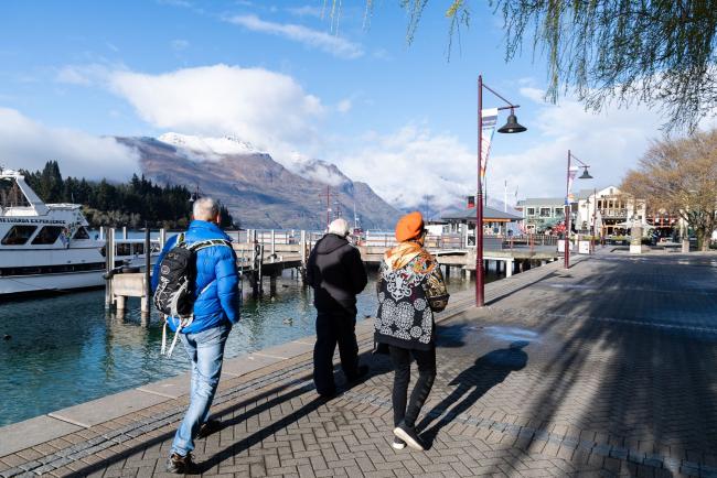 © Bloomberg. Pedestrians walk along the waterfront at Lake Wakatipu in Queenstown, New Zealand, on Thursday, Sept. 10, 2020. In a normal year, international tourism is New Zealand’s biggest export — and with its majestic alpine scenery, adventure sports and vineyards, Queenstown is usually booming. But with the country’s border closed since March 19, there are fewer foreign accents to be heard in the resort town. Photographer: Mark Coote/Bloomberg