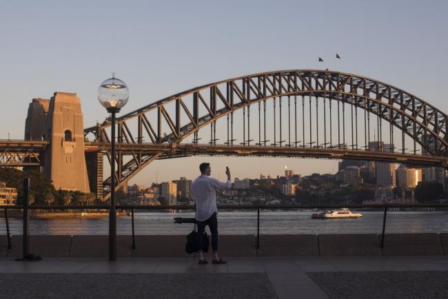 © Bloomberg. A man takes a photograph near the Sydney Harbour Bridge in Sydney, Australia, on Tuesday, Oct. 13, 2020. Australia last week released a fiscal blueprint that pushes debt and deficit to a peacetime record just hours after the central bank signaled a willingness to ease further as both arms of policy press to drive down unemployment. Photographer: Brent Lewin/Bloomberg