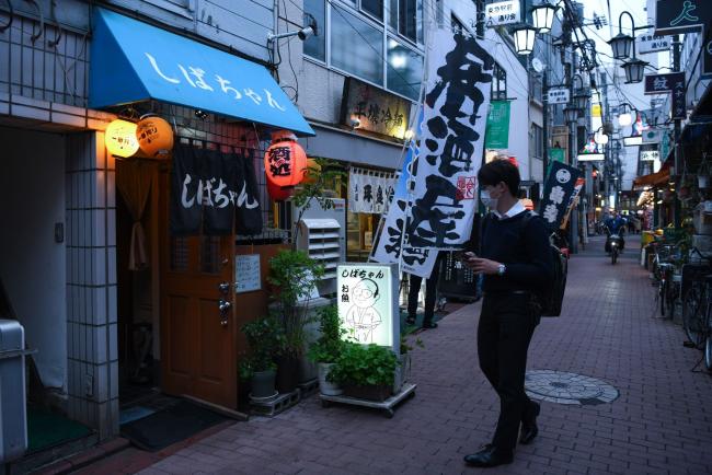 © Bloomberg. A pedestrian wearing protective face masks walk past a restaurant at night in the Kamata district of Tokyo, Japan, on May 22, 2020. The Bank of Japan launched a new lending program worth 30 trillion yen ($279 billion) to support small businesses struggling amid the coronavirus but held off from adding major stimulus at an emergency meeting Friday. Photographer: Noriko Hayashi/Bloomberg