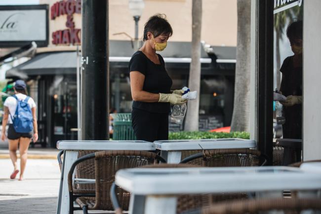 © Bloomberg. An employee wearing a protective mask and gloves at sanitizes a table in the outdoor dining area of a restaurant in Miami, Florida, U.S., on Wednesday, July 8, 2020. A day after announcing pending closures, Mayor Gimenez said he had met with medical experts and business leaders and decided that restaurants could still provide outdoor dining and gyms could open if everyone wore a mask.