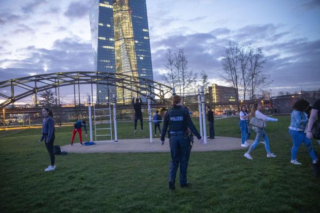 © Bloomberg. A police officer disperses people gathered at a park during social distancing operations to control the spread of coronavirus near the European Central Bank (ECB) headquarters at night in Frankfurt, Germany, on Wednesday, March 18, 2020. Europe’s bonds looked set for gains after the European Central Bank came to the rescue of debt markets for a second time this month with a 750 billion euro ($820 billion) quantitative easing package. Photographer: Alex Kraus/Bloomberg