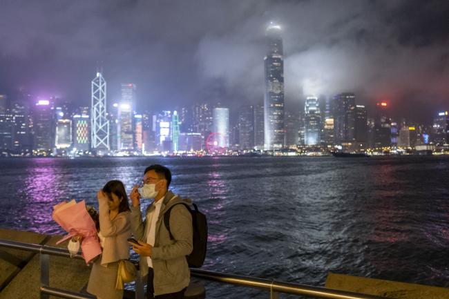 © Bloomberg. People adjust their hair and a protective mask while posing for a selfie photograph against the city skyline at night in the Tsim Sha Tsui district of Hong Kong, China, on Friday, Feb. 14, 2020. As fears of the novel coronavirus (Covid-19) spread across the region, pharmacies and supermarkets in Hong Kong are running out of basic supplies like toilet paper, paper towels, hand sanitizer and especially masks. Photographer: Justin Chin/Bloomberg